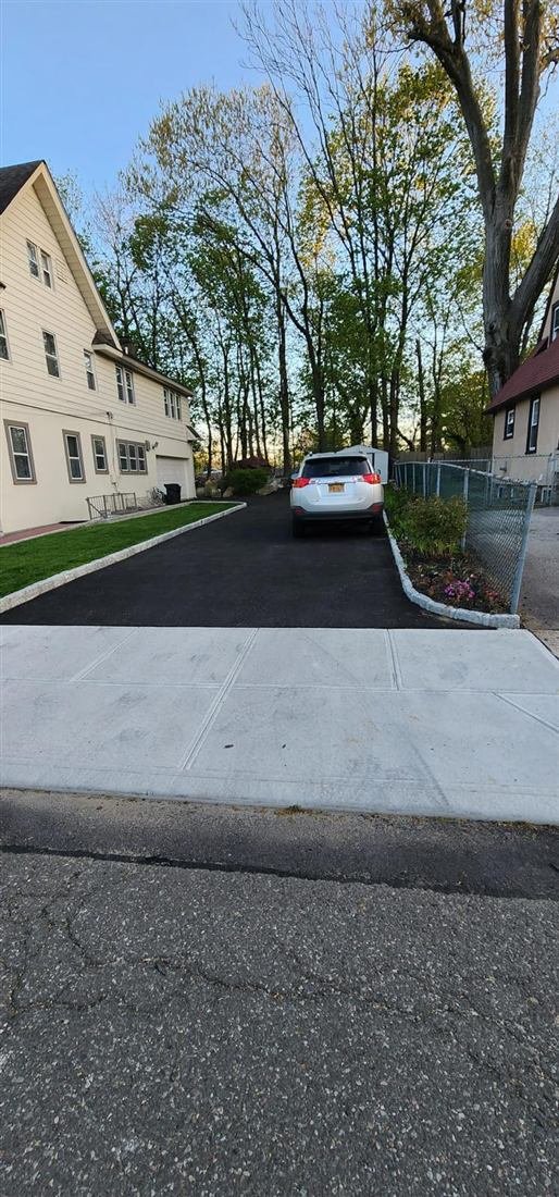 Clean photo of a completed residential blacktop asphalt driveway with Belgian block edging, new concrete sidewalk apron, fresh sod, and planted border along a chain-link fence, shot in soft evening light.