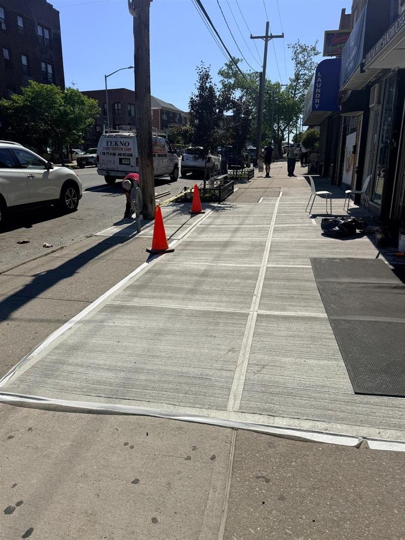 Freshly poured and taped-off commercial sidewalk replacement in front of NYC storefronts (laundry/bodega strip) with cones, Tekno Construction Corp branded van and a crouching worker at the curb, pedestrians in the distance.